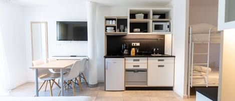 Una kitchenette moderna y compacta con microondas, horno y cafetera integrados. Cuenta con un pequeño comedor con mesa y sillas, y un televisor montado en la pared.