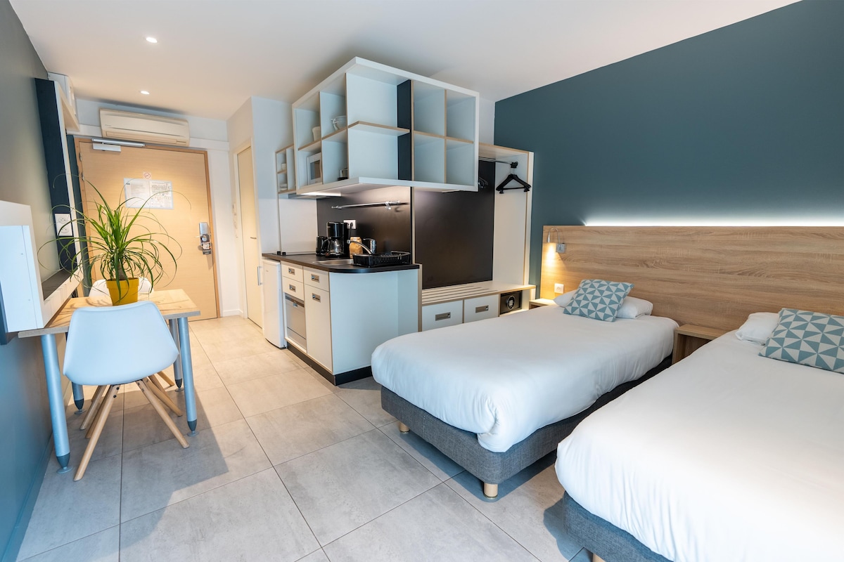 A modern hotel room with a kitchenette, a dining area, and two beds.