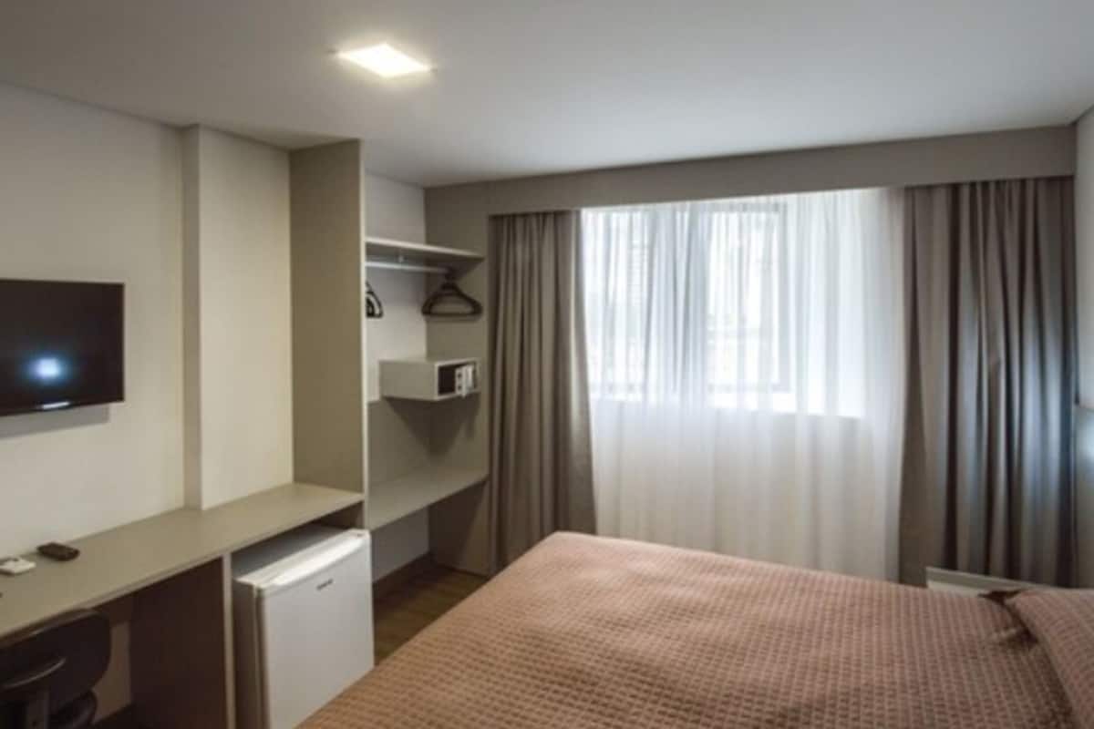Une chambre d’hôtel avec un lit, un bureau, une télévision et une fenêtre avec des rideaux.