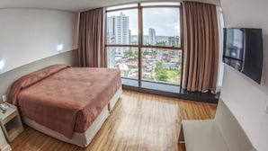 A hotel room with a large window, a bed, a bedside table, a TV, and a wooden floor. - Marante Executive Hotel (Recife)