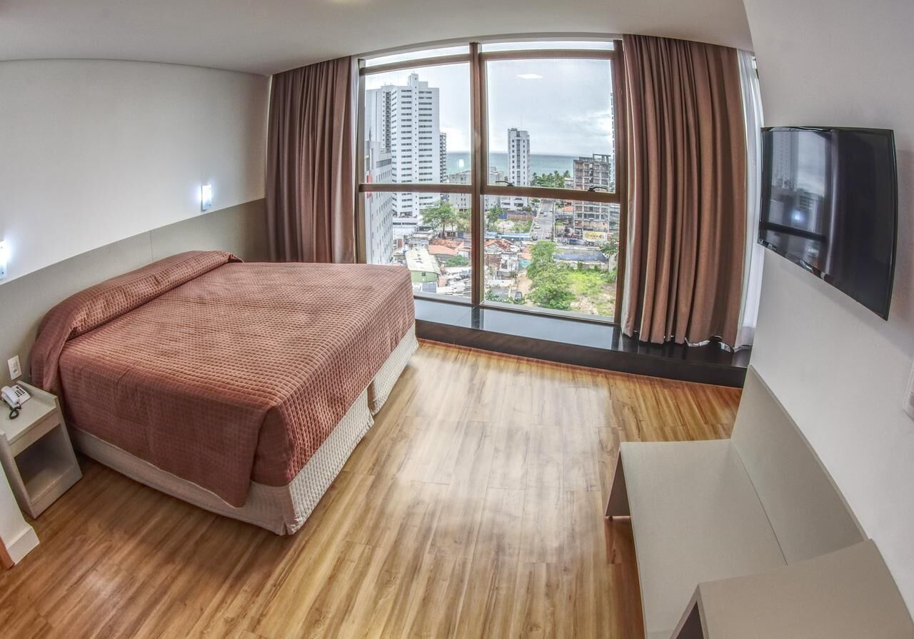 A hotel room with a bed, a nightstand, a TV, and a large window with a city view.