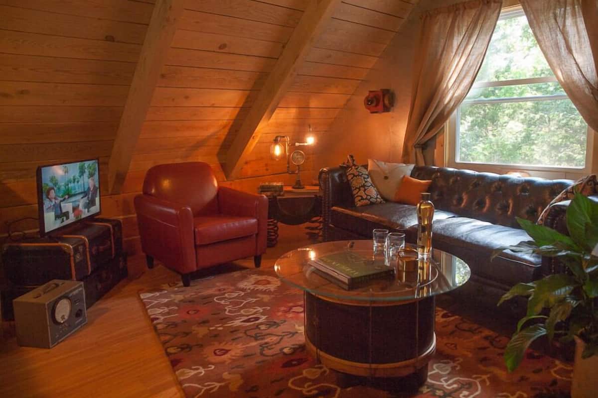 Um quarto de sótão acolhedor, com teto de madeira, um sofá de couro, uma poltrona vermelha, uma mesa de centro de vidro e uma televisão sobre um suporte de madeira.