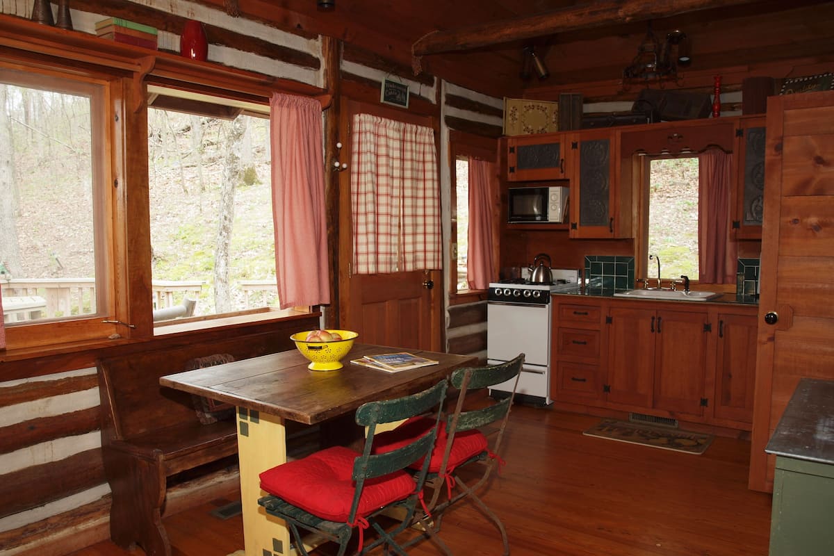 Interior rústico de cabana com mesa de jantar de madeira, cadeiras, fogão e pequena cozinha.