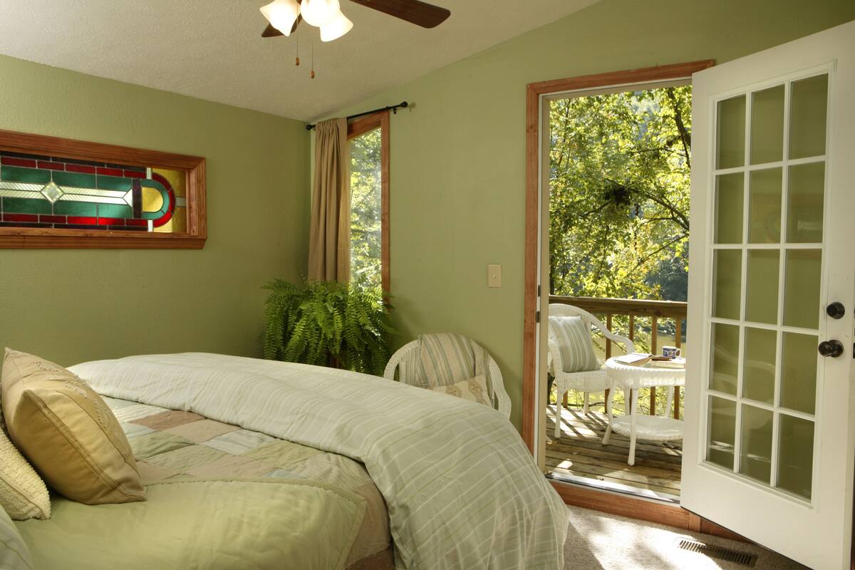 Quarto com cama, janela com cortinas, porta que dá para uma varanda e uma janela de vitral na parede.