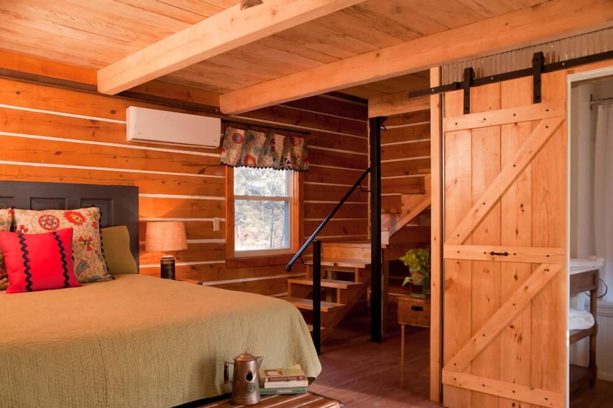 Quarto rústico em cabana de madeira, com cama, almofadas, abajur, cafeteira e porta de celeiro deslizante.