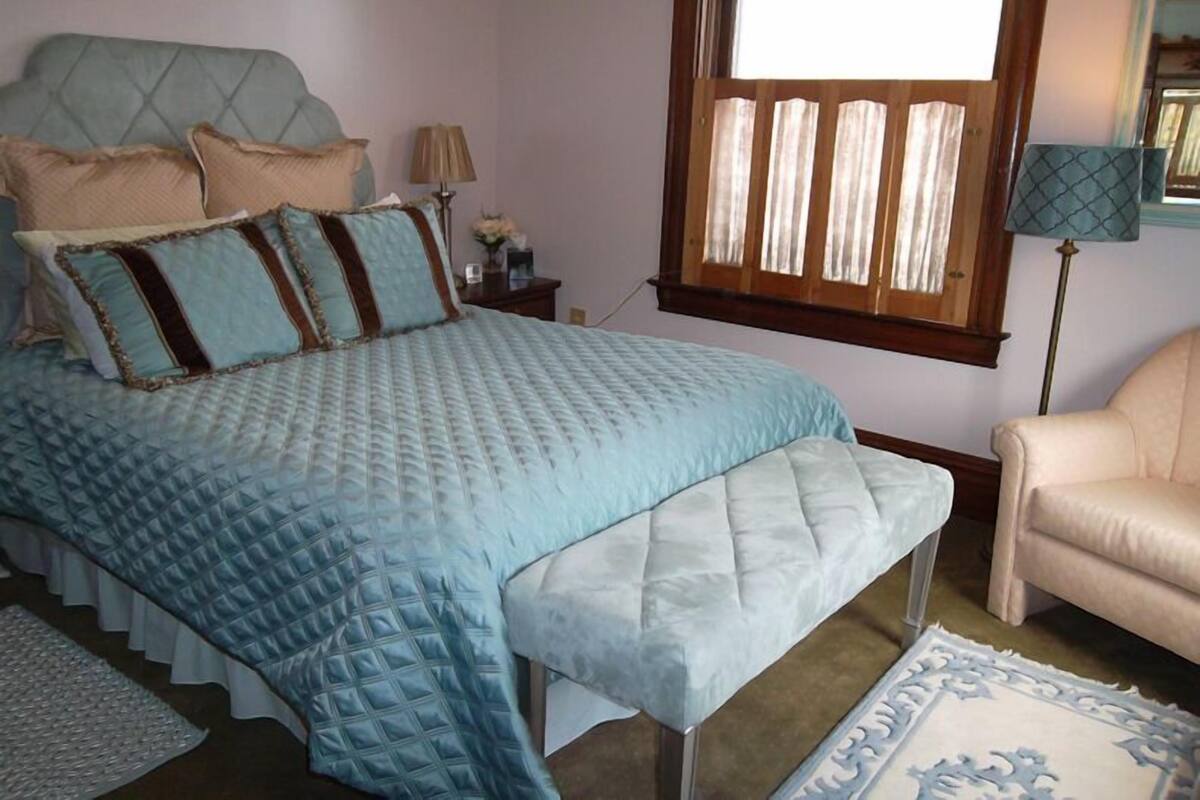 A bedroom with a bed, a bench, a chair, a nightstand, a lamp, and a window with curtains.