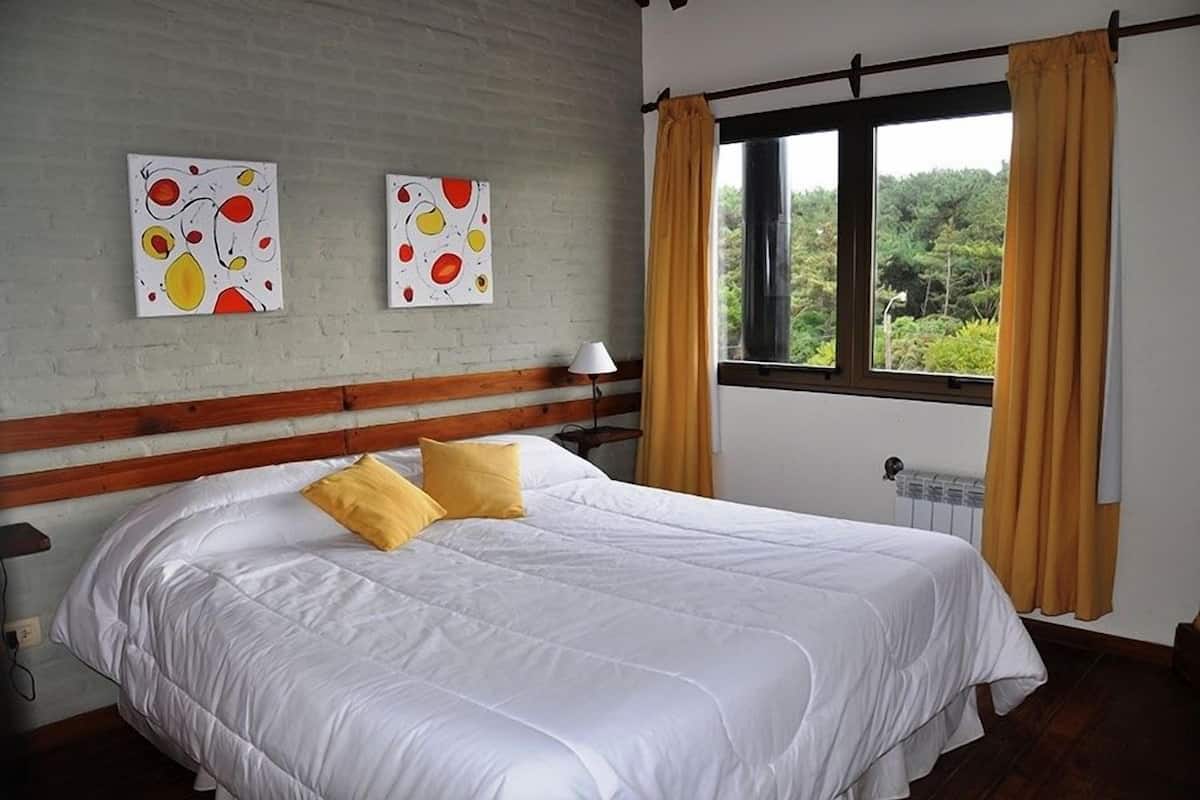 A bedroom with a large bed, two paintings on the wall, a window with curtains, and a radiator.