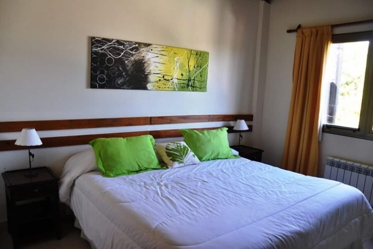 A bedroom with a bed, green pillows, a nightstand with a lamp, a painting on the wall, and a window with curtains.