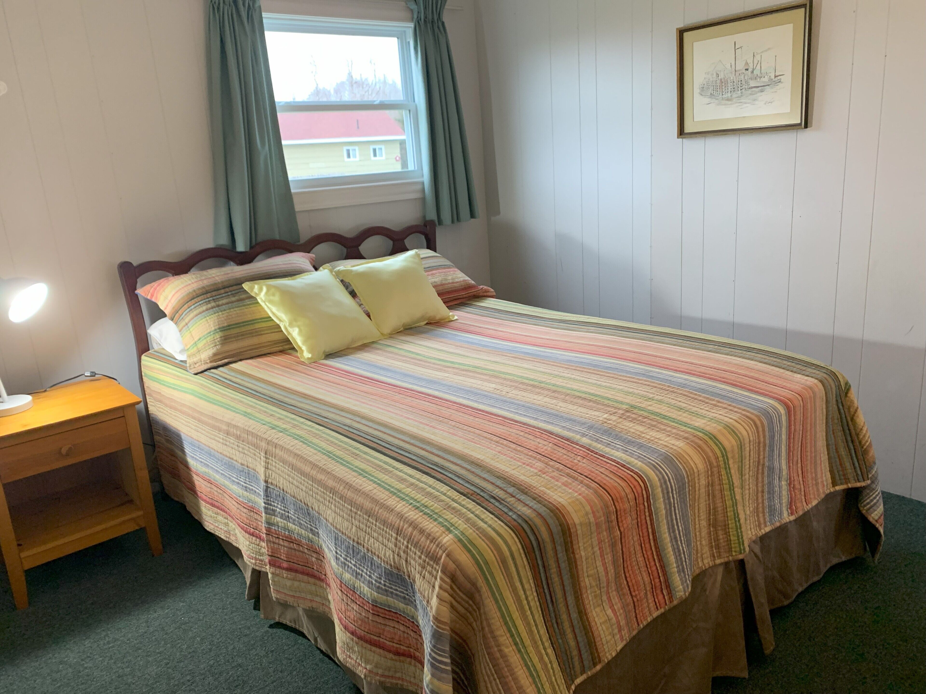 A neatly made bed with a striped bedspread, a wooden nightstand with a lamp, and a window with curtains.