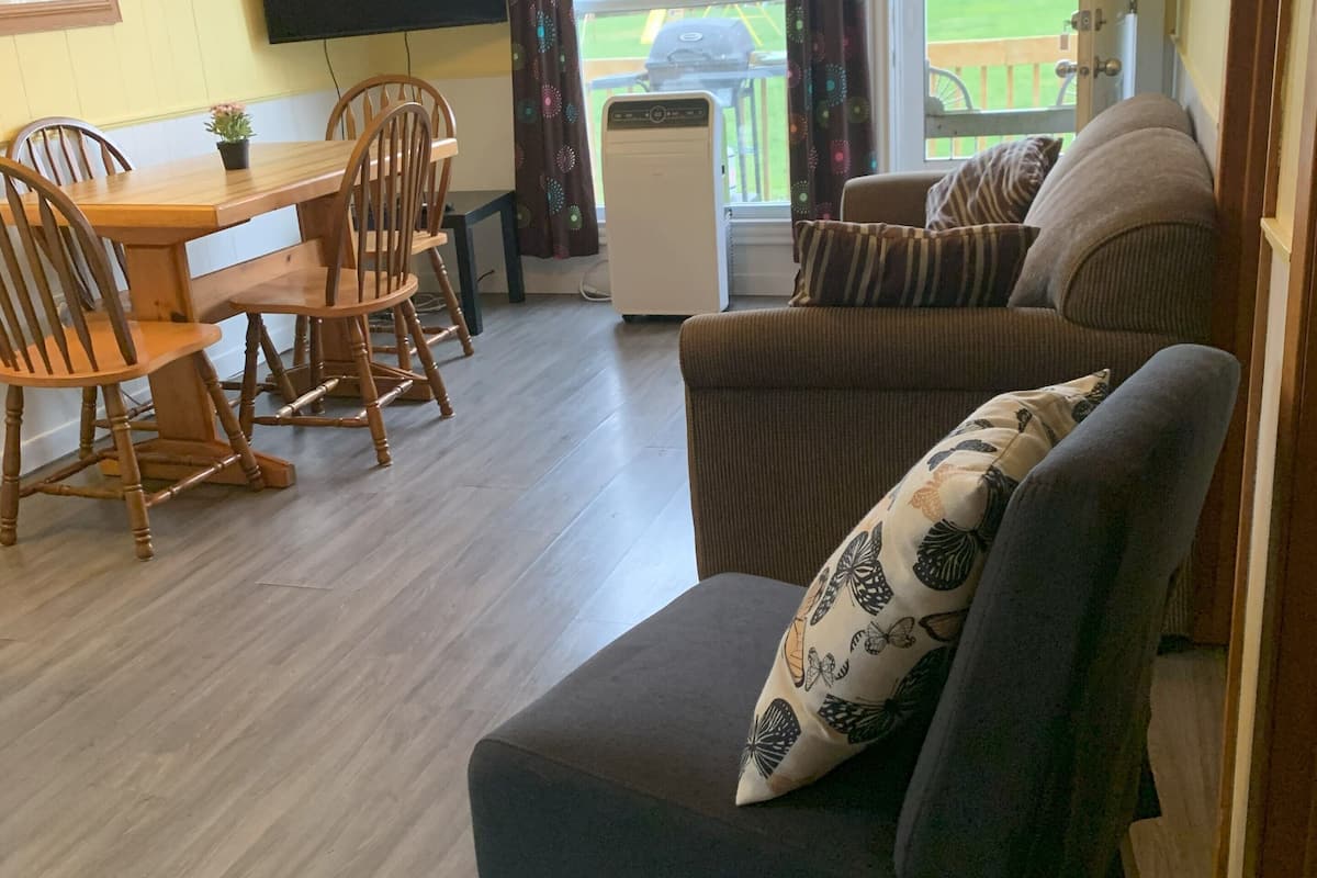 A living room with a sofa, a dining table with chairs, a TV mounted on the wall, and a window with curtains.