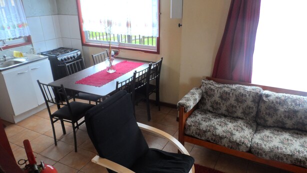 A living room with a sofa, a dining table with chairs, a kitchen area with a stove, and a window with a view of the outside. - Cabañas ROHE (Puerto Montt)