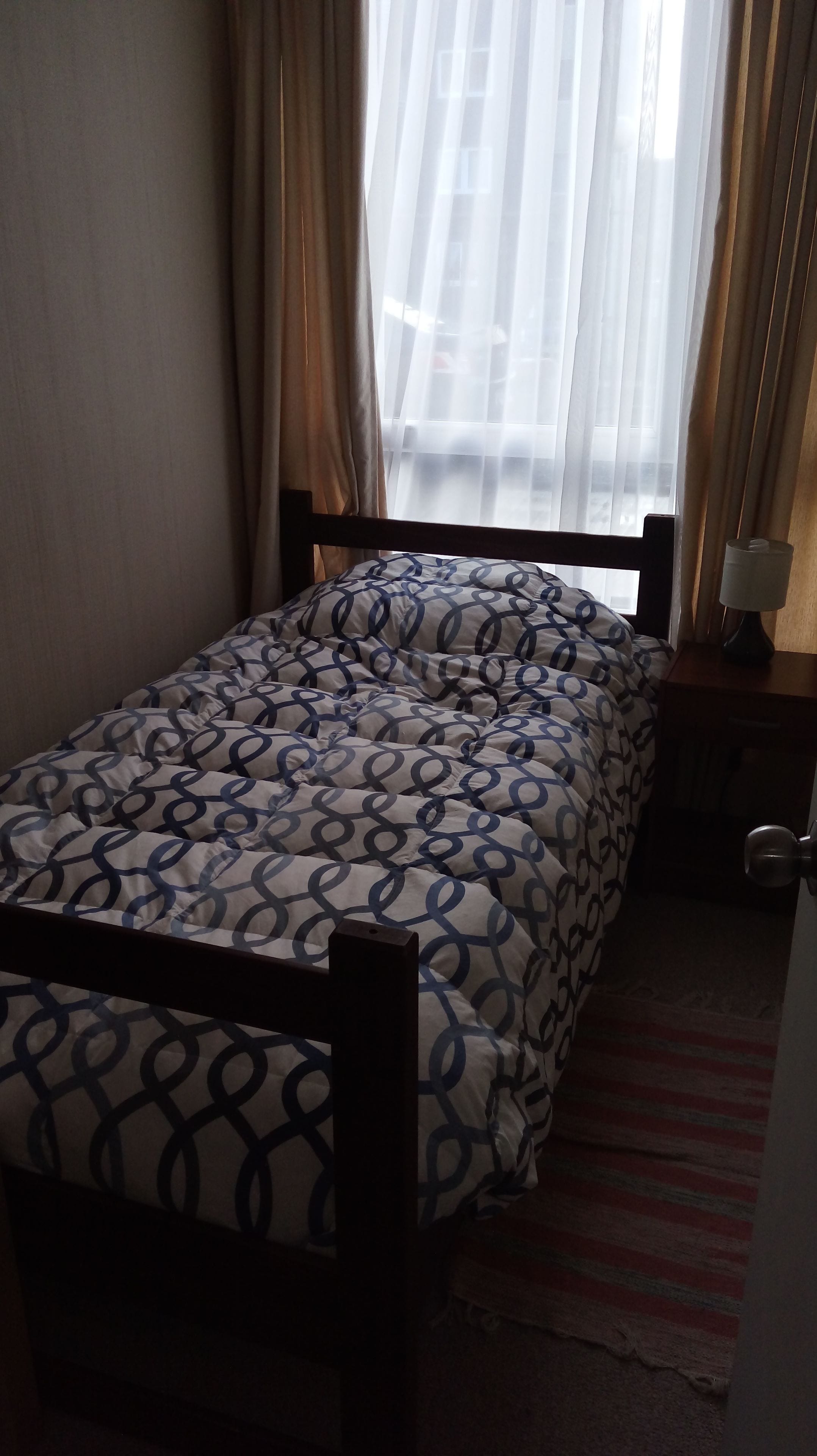A bed with a patterned bedspread, a wooden headboard, a nightstand with a lamp, and a window with sheer curtains.