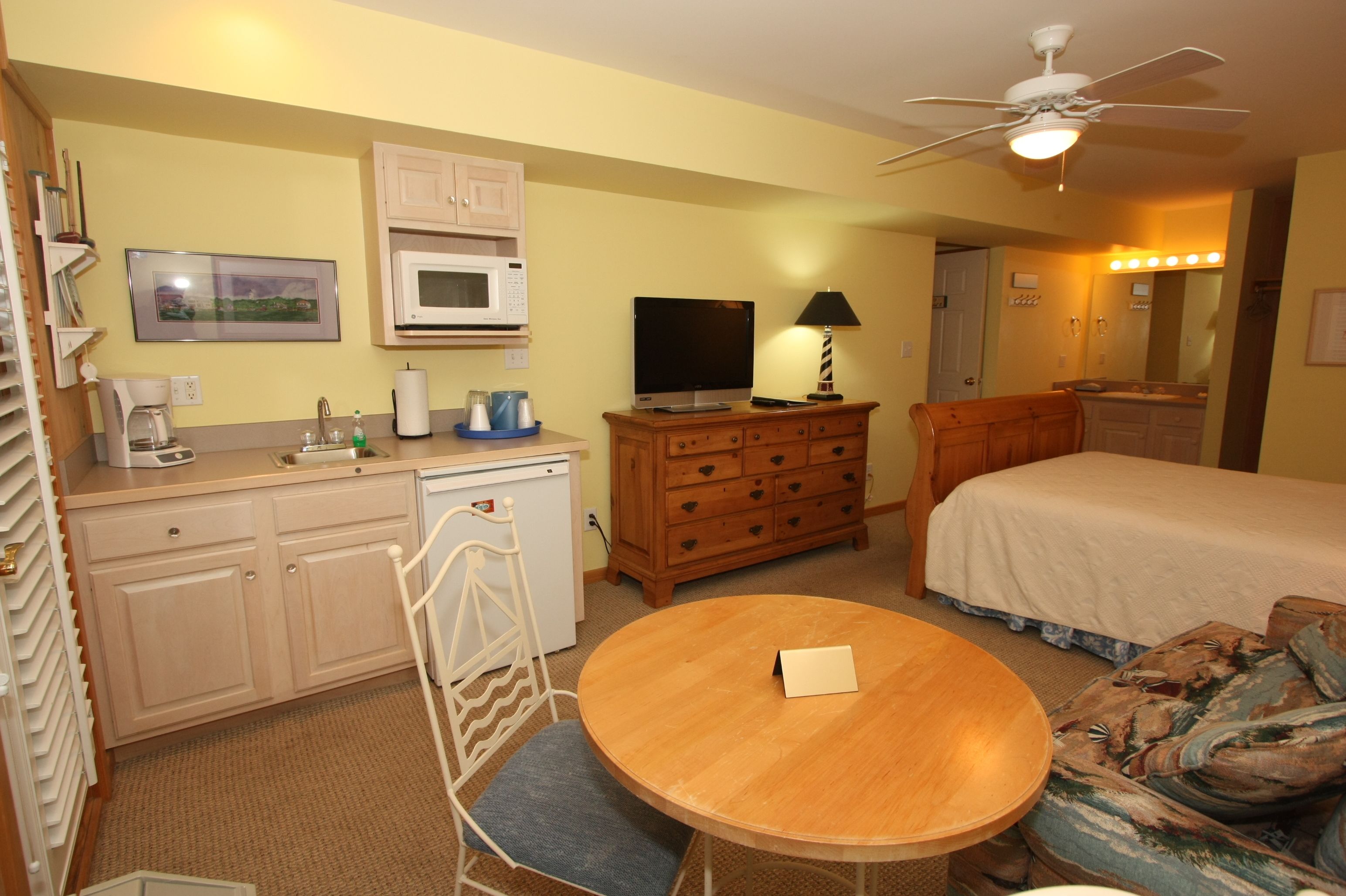 A hotel room with a bed, a desk with a microwave, a television, a wooden dresser, a ceiling fan, and a small round table.
