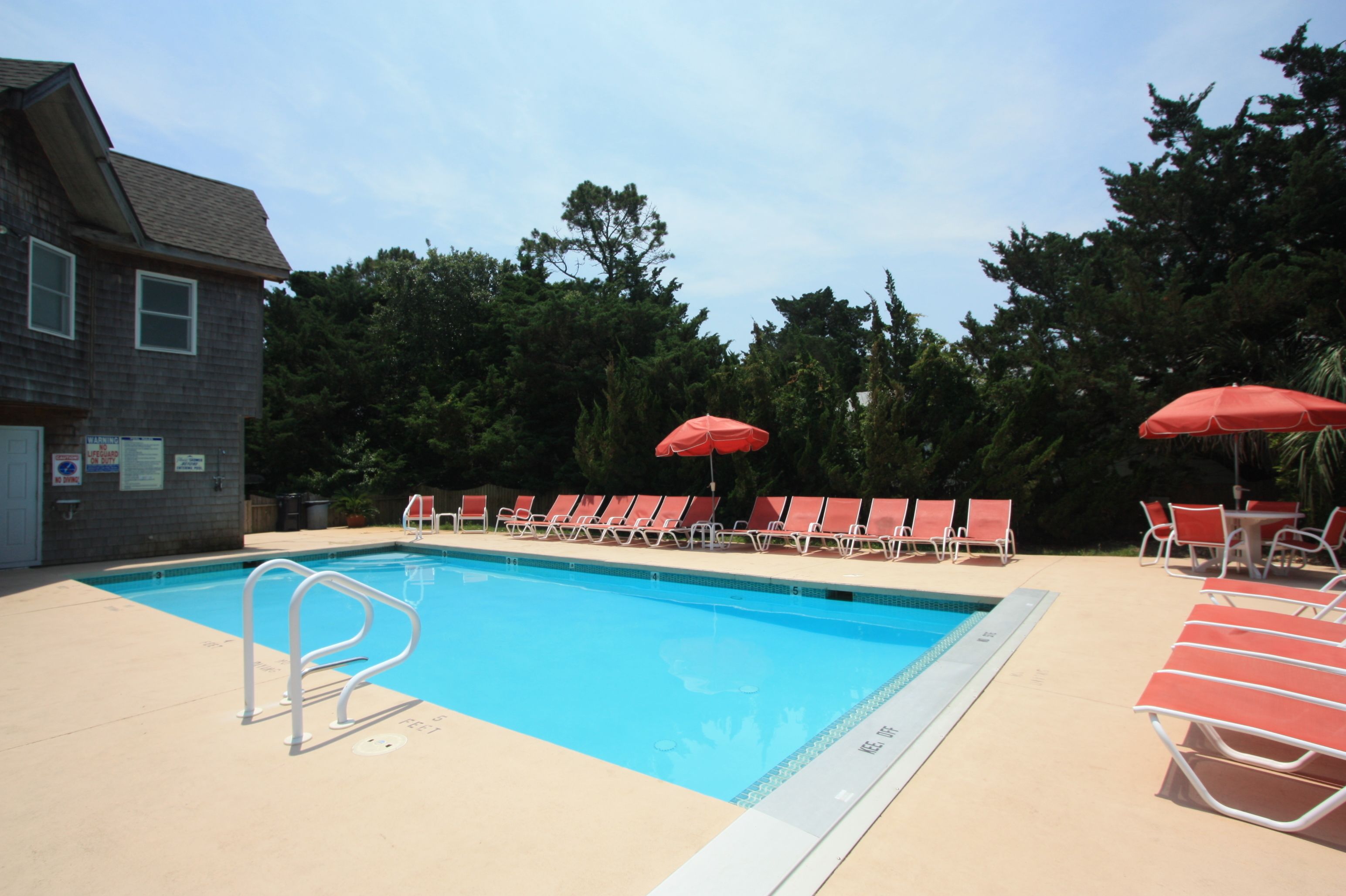 Piscina con sillas de descanso rojas y sombrillas, rodeada de árboles y un edificio.