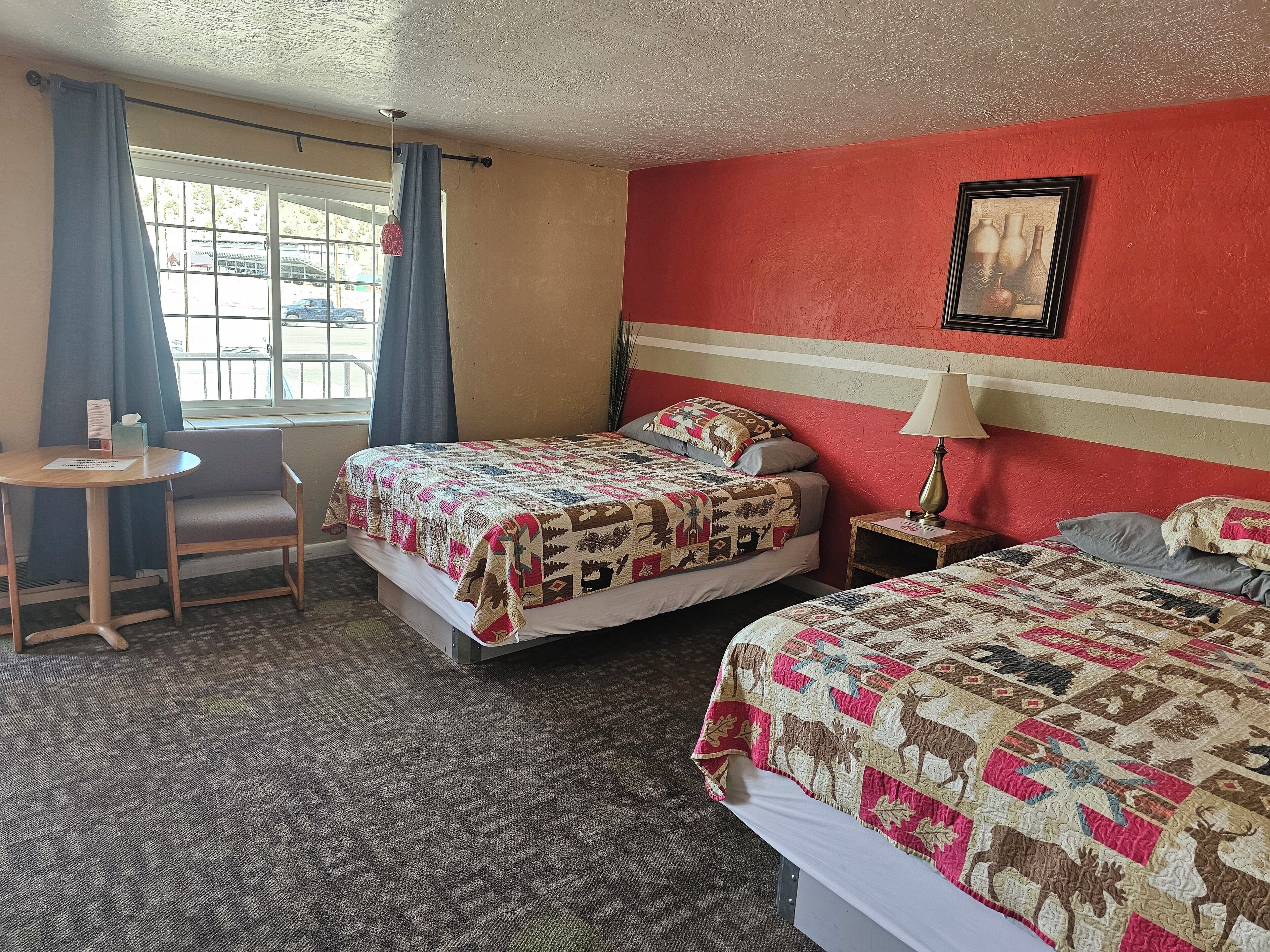 A hotel room with two beds, a small table, a chair, and a window with curtains.