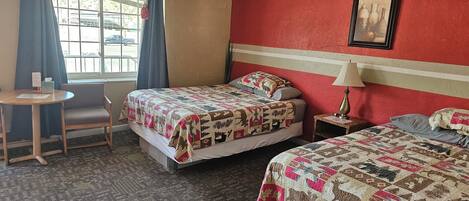 A hotel room with two beds, a small table, a chair, and a window with curtains.