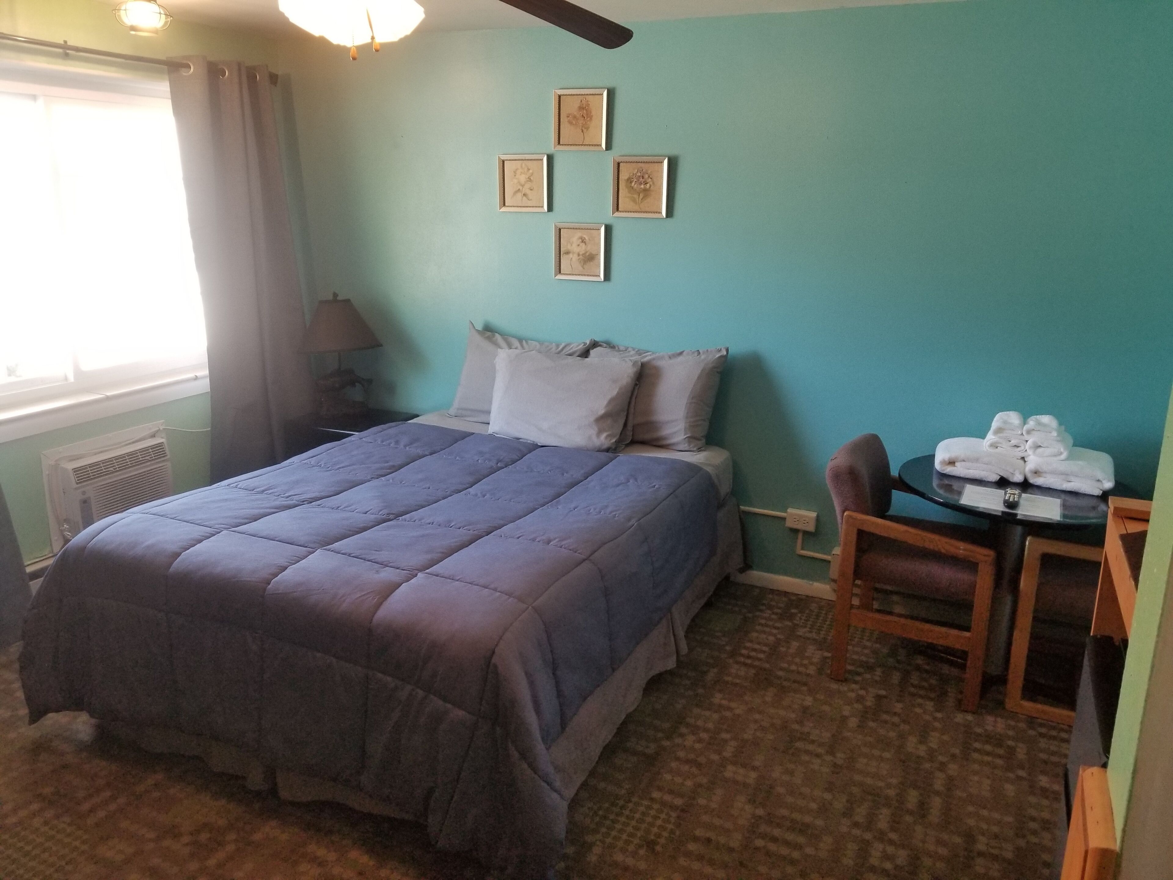 A neatly arranged hotel room with a bed, a small table with towels, a chair, and framed pictures on the wall.