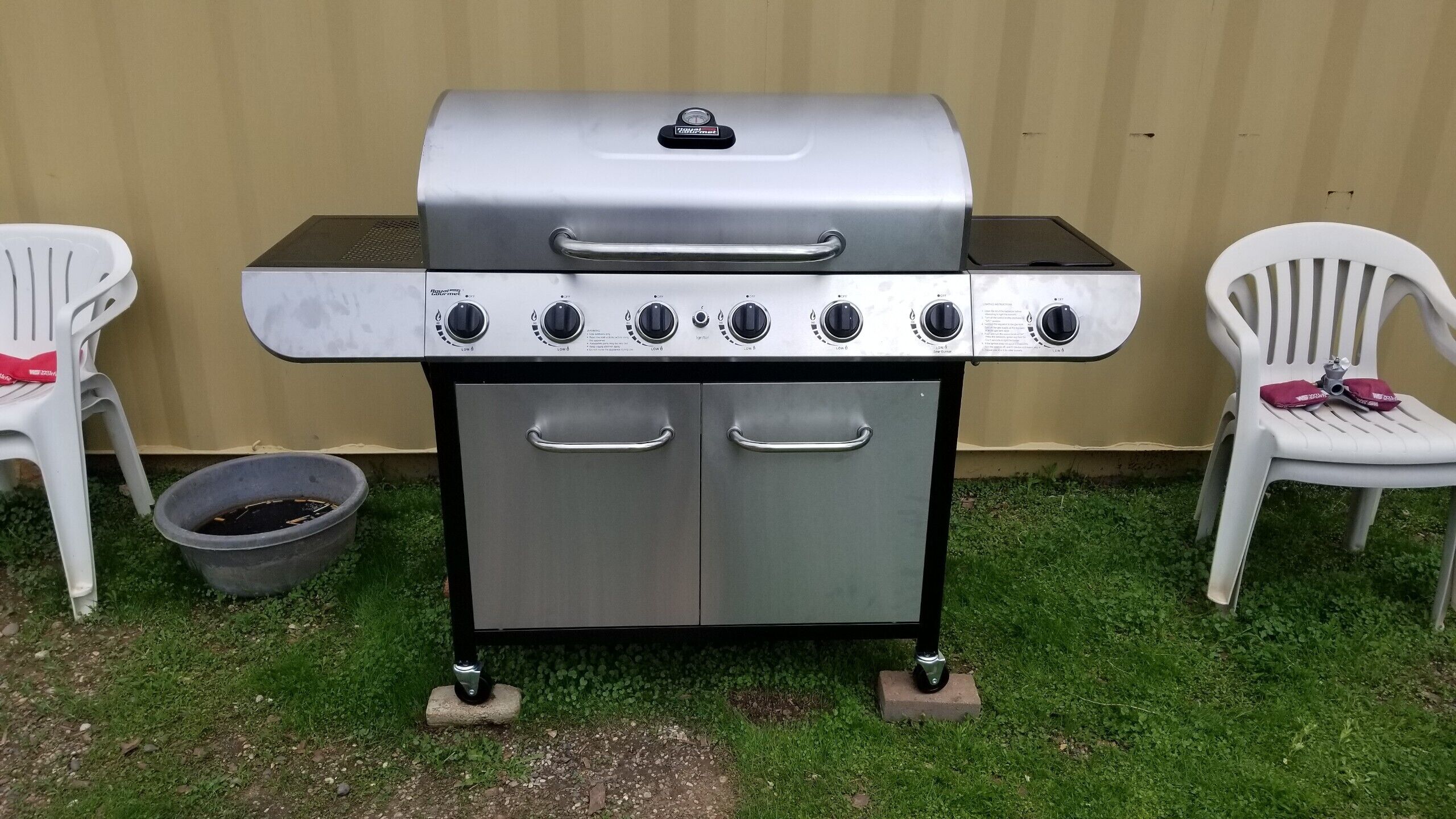 A stainless steel barbecue grill with four burners and a side shelf, placed on a grassy area with two white plastic chairs nearby.