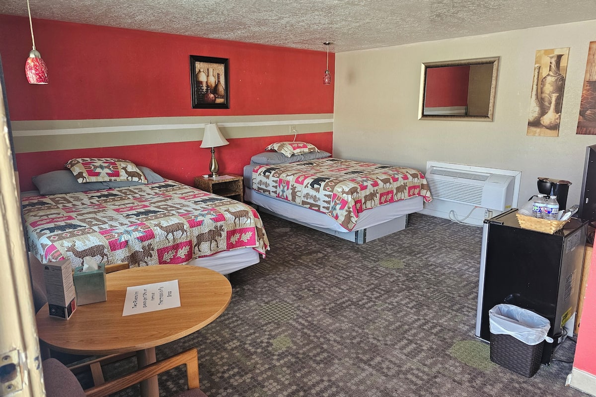 A hotel room with two beds, a small table, a desk with a computer, and a radiator.