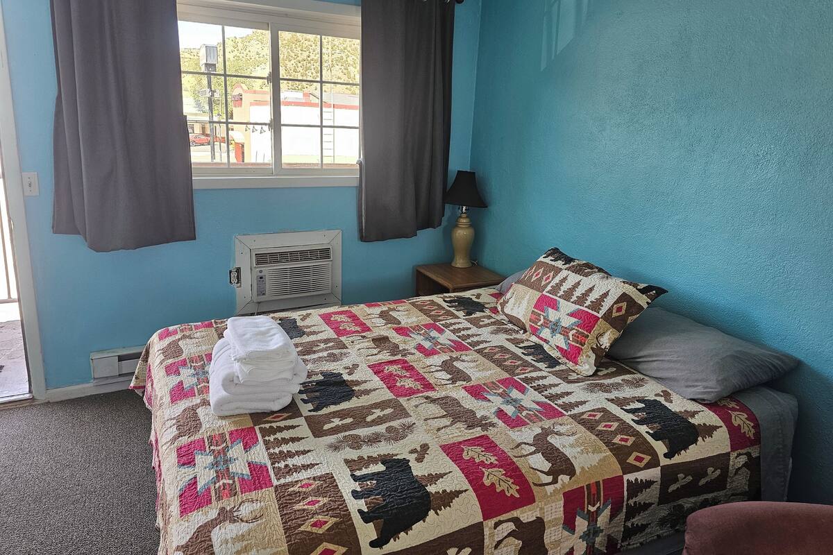 A bedroom with a blue wall, a bed with a quilt, a window with curtains, a bedside lamp, and an air conditioner.