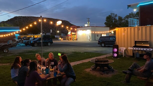 An outdoor gathering with people seated at a table, a barbecue grill, and a fire pit.