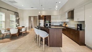 A modern kitchen with a central island, bar stools, and a dining area with a table and chairs. - Liberty Square: Private Pool, Games Room, Golf View, Near Disney (Kissimmee)