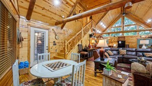 Un acogedor interior de cabaña de madera con una escalera de madera, una sala de estar con sofá de cuero, un comedor con un tablero de ajedrez y un amplio ventanal que aporta luz natural.