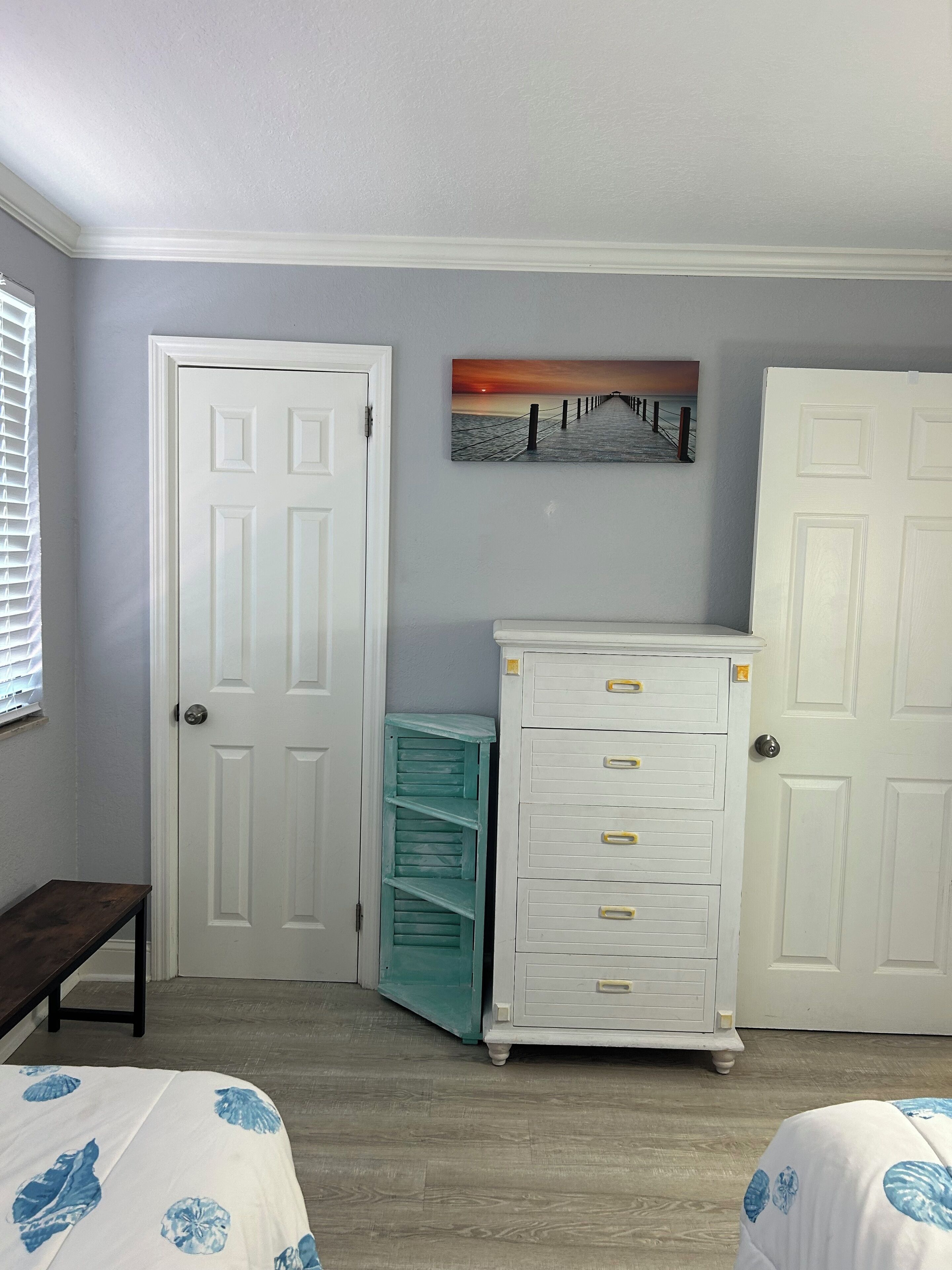 A bedroom with two white doors, a teal dresser, a white chest of drawers, a bed with a blue floral bedspread, and a sunset painting on the wall.