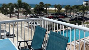 Un balcon avec une piscine, donnant sur une plage bordée de palmiers et où sont garées des voitures.