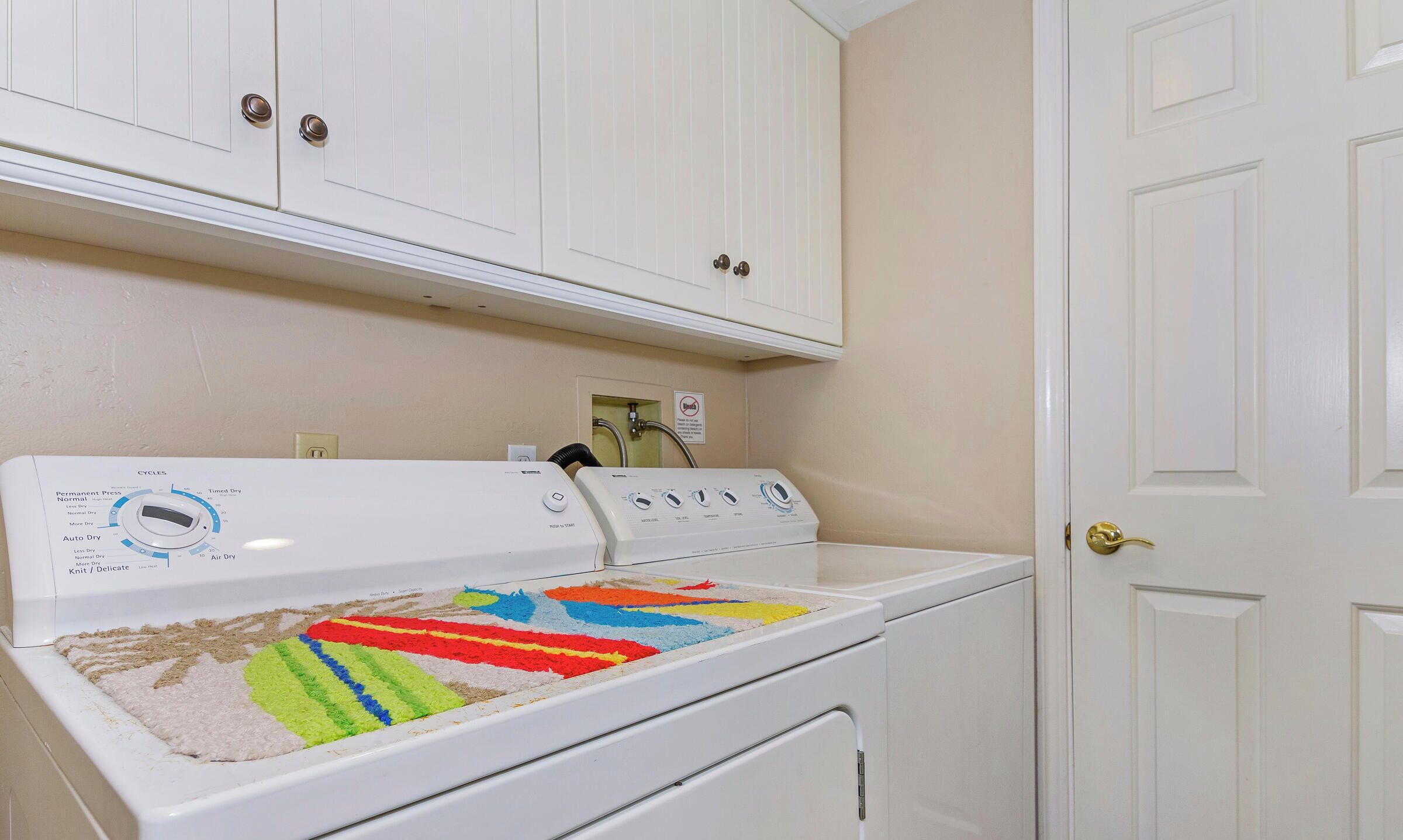 A white front-loading washer and dryer with a colorful drying rack, white cabinets above, and a white door to the right.