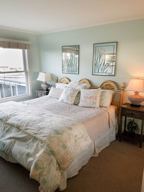 A bedroom with a large bed, two bedside tables, a window with blinds, and two wall-mounted artworks.