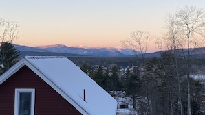 A red cabin with a snow-covered roof, surrounded by trees and mountains in the background. - 10 Min to Sunday River, Sleeps 16, Pool Heated Year-Round, Mountain Views (Bethel)