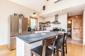 A modern kitchen with a stainless steel refrigerator, a central island with chairs, wooden cabinets, and a built-in oven. - Heated Pool in Winter, Sleeps 18, 10 Min to Sunday River, Mountain Views (Bethel)