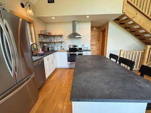 Una cucina moderna con isola centrale, elettrodomestici in acciaio inossidabile, pavimento in legno e una scala che conduce a un piano superiore.
