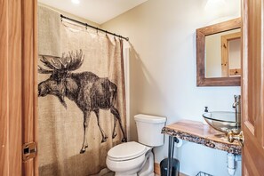 A bathroom with a moose shower curtain, a toilet, a sink with a glass basin, and a wooden-framed mirror.