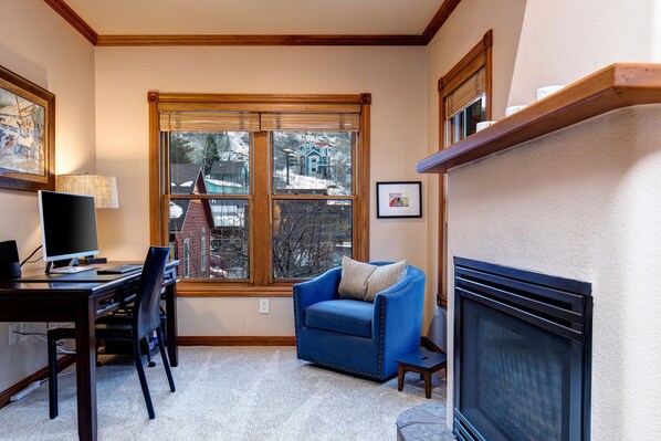 A living room with a fireplace, a desk with a computer, a blue armchair, and a large window with a view of a house.