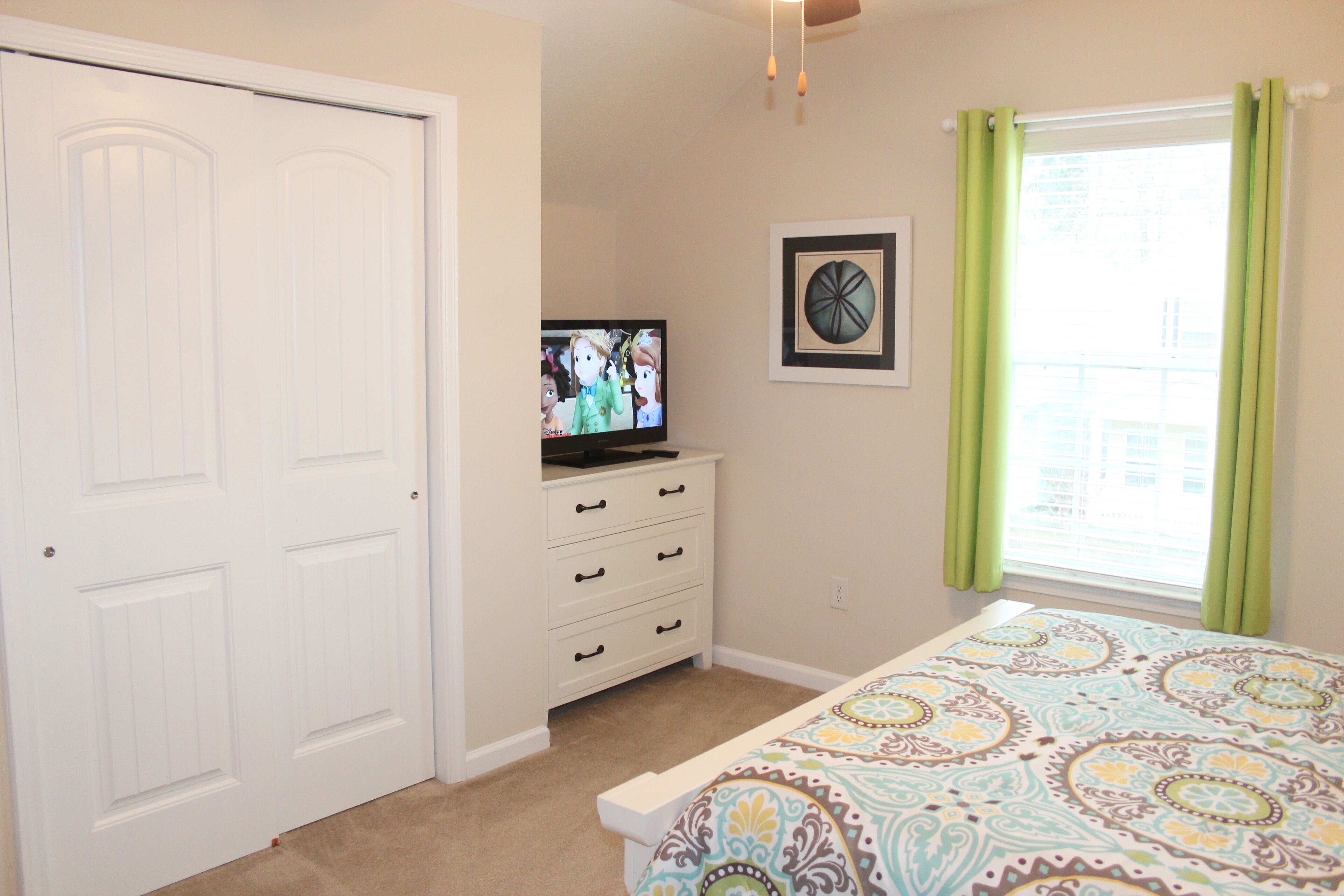 A bedroom with a bed, a dresser, a television, a window with curtains, and a door.