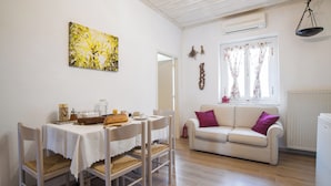 Ein Essbereich mit einem für zwei Personen gedecktem Tisch, einem weißen Sofa mit einem violetten Kissen und einem Fenster mit blumigen Vorhängen.