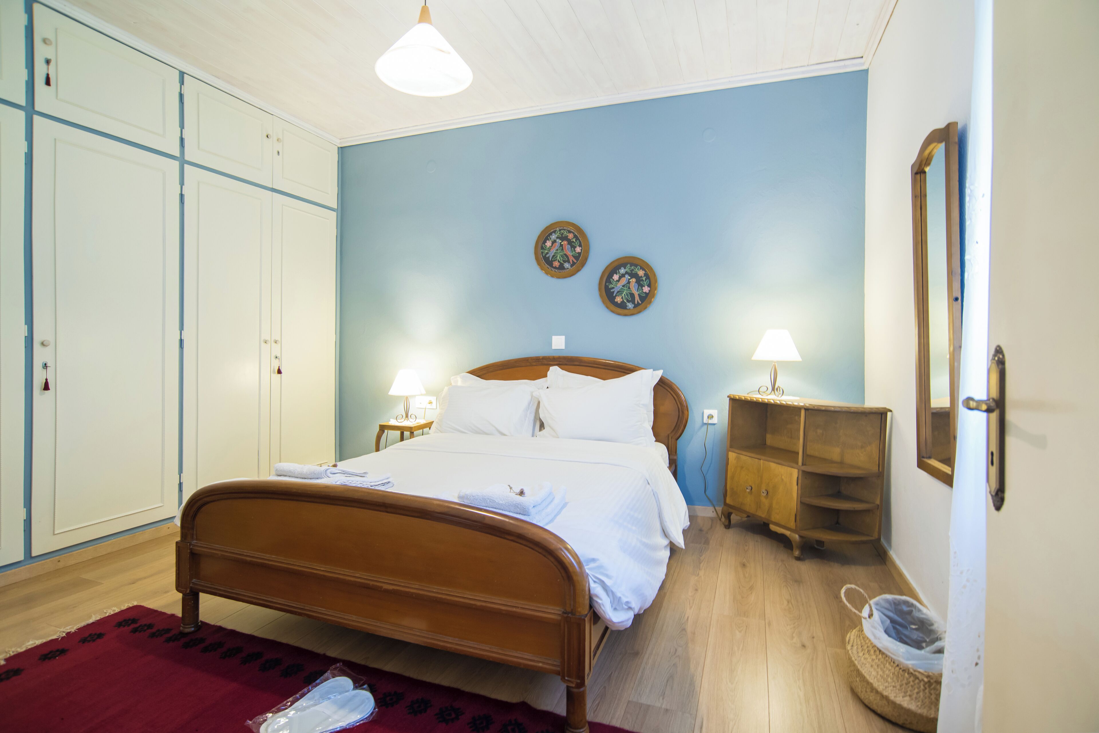 A bedroom with a wooden bed, a red rug, a wooden nightstand, a lamp, and a mirror on the wall.