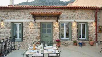 Un bâtiment en pierre avec un toit recouvert de tuiles, un coin repas avec une table dressée pour quatre personnes, et des plantes en pot sur le porche.