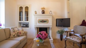 Ein Wohnzimmer mit beiger Couch, einem Holztisch mit Blumenvase, einem Kamin mit einem Gemälde, einem Fernseher und einem Holzstuhl.