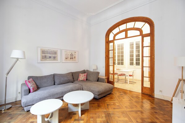 Een woonkamer met een grijze bank, twee witte tafels en een houten vloer.