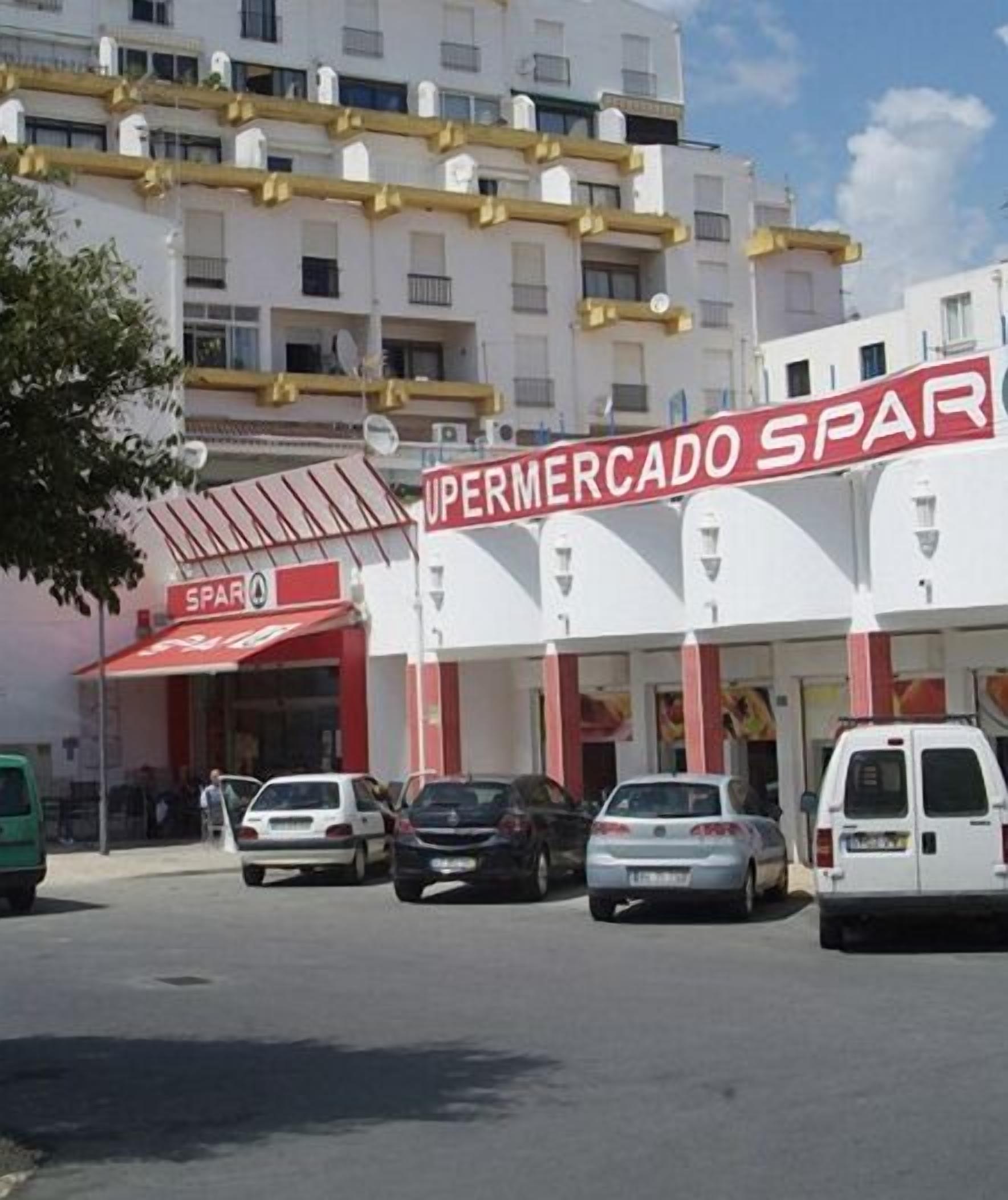 A supermarket named 'Supermercado SPAR' with a red and white facade, parked cars in front, and a multi-story building in the background.