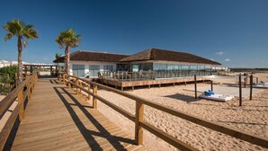 Un hotel frente al mar con un paseo marítimo de madera que conduce a un edificio con techo de vidrio, palmeras y una playa de arena.