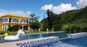 A swimming pool with a tiled edge, a yellow building with a balcony, and a lush green hillside in the background. - Rak Na Suan Pueng Resort (Suan Phueng)