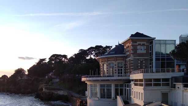 A coastal building with a balcony overlooking the beach, a pathway leading to the entrance, and a view of the sea.