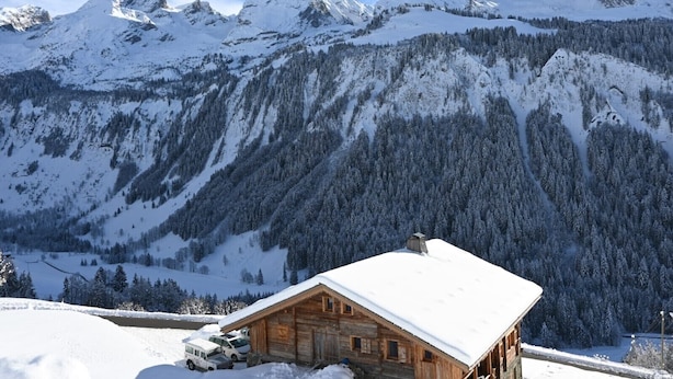 A wooden cabin with a snow-covered roof, surrounded by snow-covered mountains and trees.