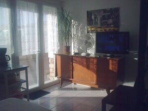 A room with a wooden cabinet, a television, a potted plant, and a large window with curtains. - Air conditioned studio close to free parking, shopings and beaches (Carqueiranne)