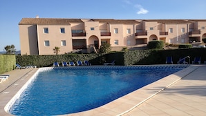 Una piscina con agua azul, rodeada de baldosas color beige. Hay sillas reclinables azules a lo largo del borde de la piscina.