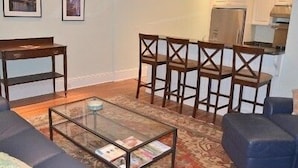 A living room with a glass coffee table, blue sofa, and a patterned rug. There are framed pictures on the wall and a kitchen area with cabinets and a refrigerator. - Fabulous 1890 Victorian home on Capitol Hill; 100 steps to metro, #1 (Washington)